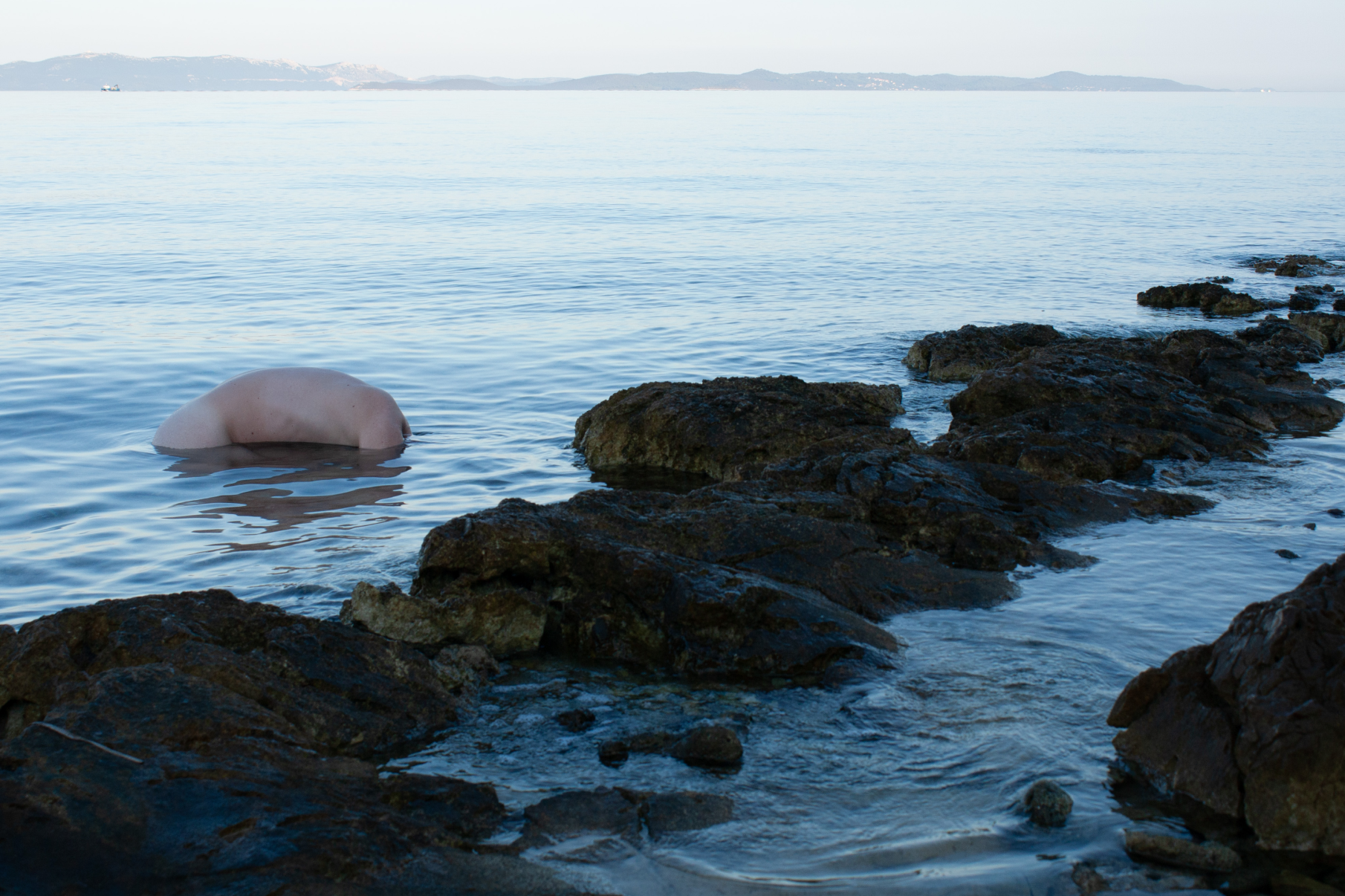 Human figure partly submerged in calm water beside a rocky coastline